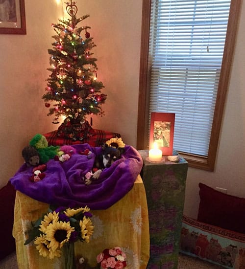 An altar with a small decorated Christmas tree, colorful cloths, and various items including framed photos, a lit candle, plush animals, sunflowers, and other flowers creates a serene space. The scene resembles a vet's thoughtful memorial corner set by a window with closed blinds.