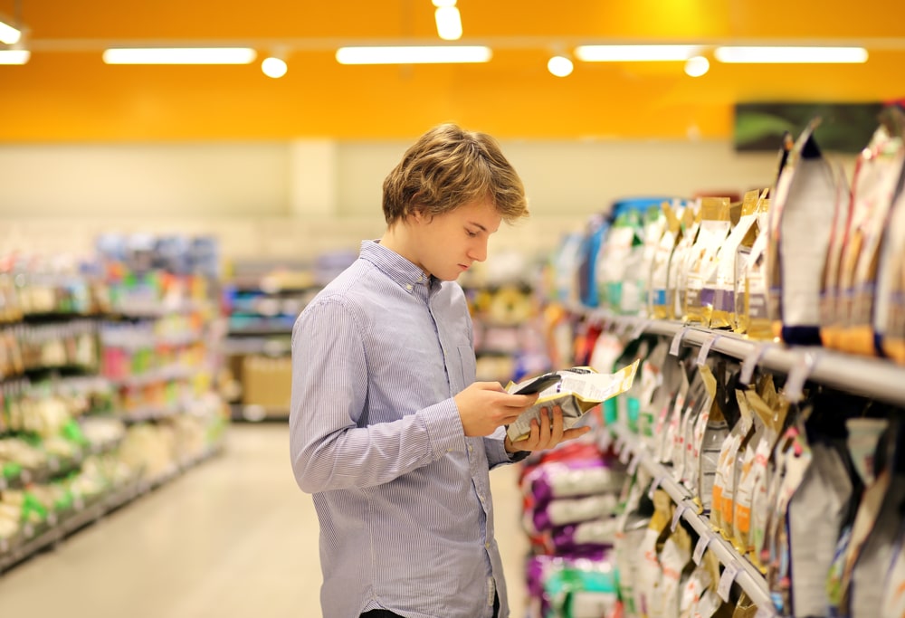 A person with light brown hair, wearing a light blue shirt, is standing in a grocery aisle like a careful veterinarian reading a product label. They are surrounded by shelves stocked with various packages, and the background is brightly lit with an orange ceiling.